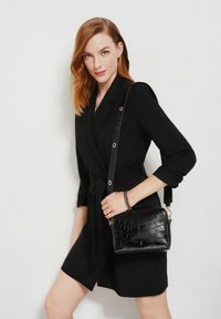 Kobieta z włosami w kolorze czerwonym do ramion, nosząca czarną sukienkę żakietową z paskiem i trzymająca czarną torbę na ramię o teksturowanej powierzchni na białym tle.