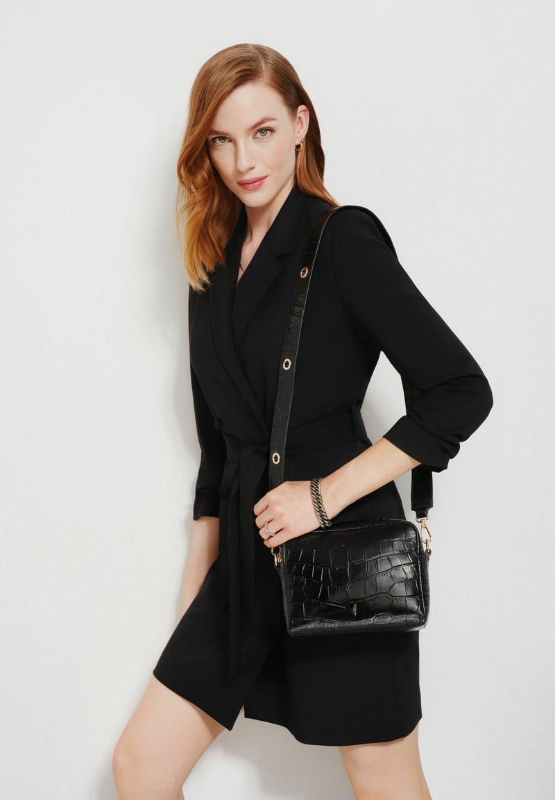 Kobieta z włosami w kolorze czerwonym do ramion, nosząca czarną sukienkę żakietową z paskiem i trzymająca czarną torbę na ramię o teksturowanej powierzchni na białym tle.