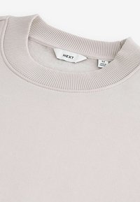 Sudadera de algodón color beige claro con cuello redondo acanalado, que presenta detalles de costuras y una etiqueta que muestra las instrucciones de cuidado y la talla (M).