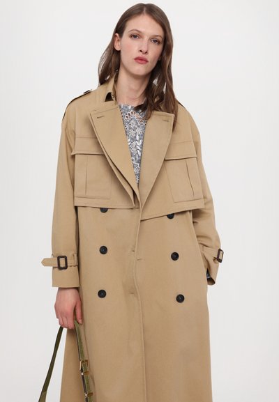 Cappotto beige con bottoni doppi, grandi tasche frontali e spalline sulle spalle. Presenta una texture morbida e maniche con cintura.