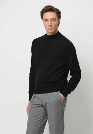 Schwarzer Rollkragenpullover aus weichem Material, mit langen Ärmeln und taillierter Passform. Kombiniert mit hellgrauen Hosen.