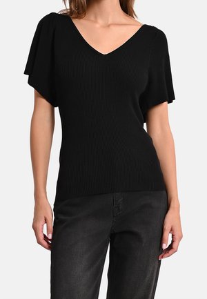 Mujer con una camiseta negra acanalada de manga corta y escote en V con mangas con vuelo, combinada con vaqueros negros, posando frente a un fondo blanco.