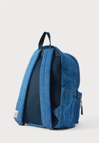 Modrý denimový batoh s polstrovaným sieťovinovým chrbtom, nastaviteľnými popruhmi, bočnou kapsou a značkovým štítkom v spodnej časti. Má zapínanie na zips.
