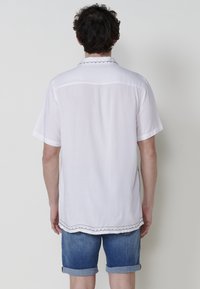 Camisa blanca de manga corta con cuello festoneado, que presenta un dobladillo recto y un sutil detalle de costura, combinada con pantalones cortos de mezclilla con puños.