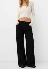 Femme portant un pull court crème et un jean noir taille haute à jambes larges, les mains dans les poches, debout devant un fond blanc.