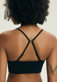 Žena s kudrnatými vlasy, která nosí černý crop top s tenkými ramínky, ukazuje záda a tetování víly na horní části pravé paže.