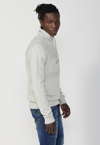 Hellgrauer Pullover mit hohem Kragen und vorderem Zugband. Mit strukturierten horizontalen Linien über der Brust und gerippten Bündchen.