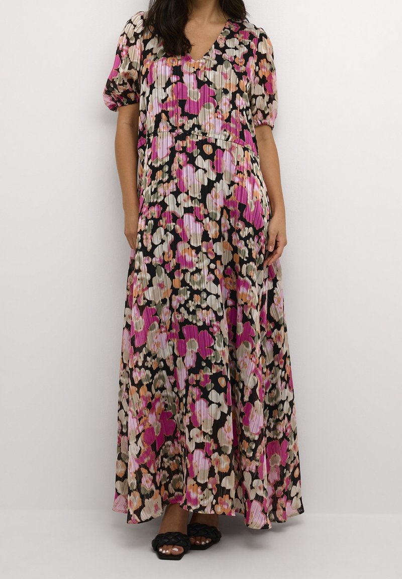 Femme portant une robe maxi plissée à manches courtes avec un motif floral rose, pêche et beige sur fond noir, et des sandales noires tressées.