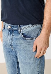 Hombre vistiendo jeans de mezclilla azul claro y una camisa de color azul marino oscuro, con la mano reposando al lado del bolsillo del jeans.