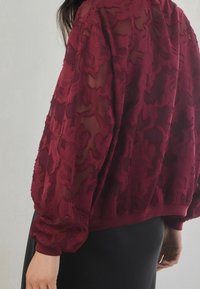 Veste texturée bordeaux avec une couche transparente ornée de motifs floraux en relief, manches ballon et taille cintrée. Vêtement noir en dessous.