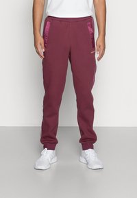 Man som bär vinröda Adidas sweatpants med satinfickor och vita sneakers står mot en tom vägg och golv.