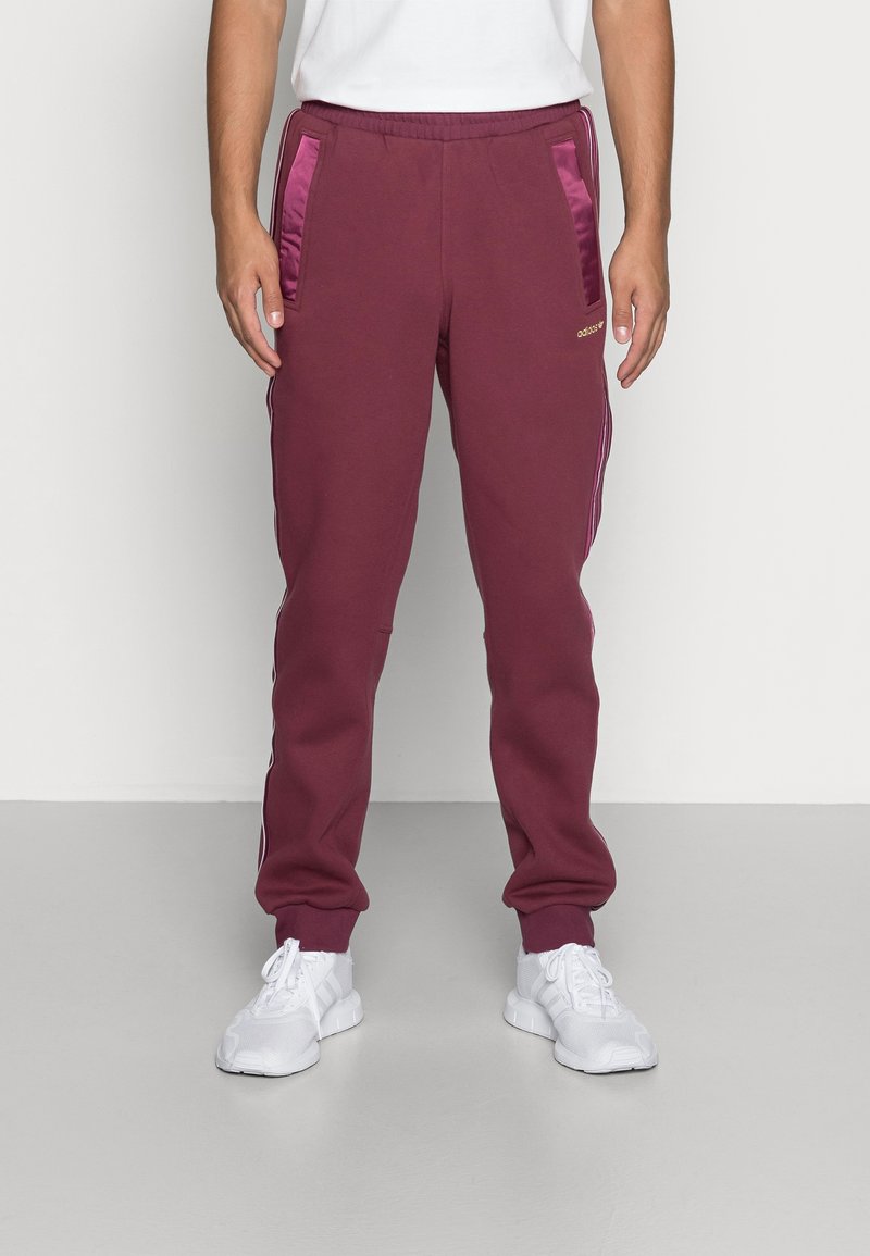 Man som bär vinröda Adidas sweatpants med satinfickor och vita sneakers står mot en tom vägg och golv.