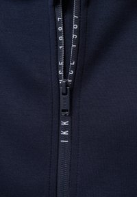 Felpa con zip di colore blu navy, dotata di una robusta cerniera con tirante nero opaco, lettering del marchio bianco a contrasto lungo i bordi, tessuto dalla superficie liscia.
