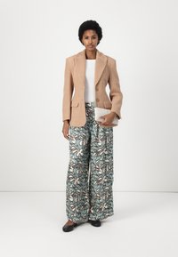 Blazer beige con texture, top bianco e pantaloni larghi con motivo in teal, crema e nero. Tiene una clutch crema con texture e ballerine nere.
