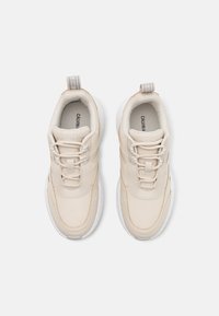 Pár béžových sneakers značky Calvin Klein Jeans s bílými podrážkami, pohled zepředu, zavázané, na světle šedém pozadí.