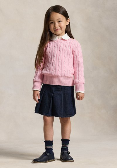 Jeune fille aux longs cheveux portant un pull rose à torsades, une chemise blanche colletée, une jupe plissée bleu marine, des chaussettes à motifs et des chaussures noires.