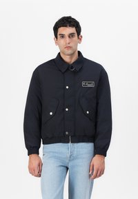 Navyblaue, kurz geschnittene Jacke mit Kragen, elastischem Bund, zwei Fronttaschen und Druckknopfverschluss. Mit einem Logo-Patch versehen.