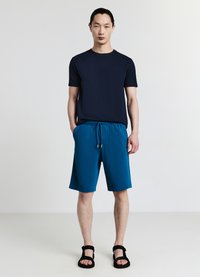 Maglietta blu navy a maniche corte con scollo a girocollo abbinata a pantaloncini turchesi con coulisse. Il modello indossa sandali neri, in piedi davanti a uno sfondo semplice.