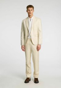 Ljust beige kostym bestående av en kavaj och byxor med elastisk midja, tillsammans med en vit skjorta och bruna skor. Enkel, skräddarsydd design.