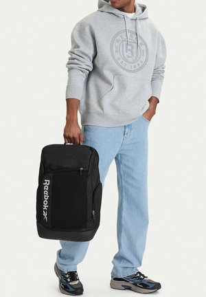 Persona con una sudadera con capucha gris de Reebok y jeans azul claro sosteniendo una mochila negra de Reebok por la asa superior.