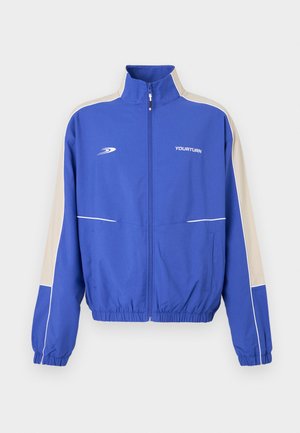 Veste de survêtement bleue zippée avec bandes latérales beiges, poignets et ourlet élastiques, et logo "YOURTURN" sur la poitrine.