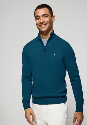 Türkiser Reißverschluss-Pullover aus strukturiertem Stoff, mit gerippten Bündchen und Saum sowie einem kleinen Logo auf der linken Brust. Das Modell trägt weiße Hosen.