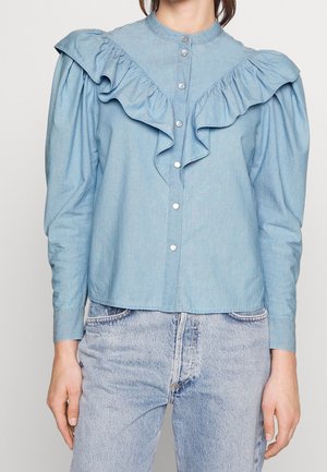 Jasnoniebieska denimowa bluzka z marszczonym przodem, zapięciem na guziki, bufkami na ramionach i długimi rękawami, zestawiona z jasnoniebieskimi jeansami o spranym wyglądzie.