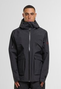 Giacca nera impermeabile con colletto alto e cappuccio regolabile, dotata di zip centrale, diverse tasche e tessuto testurizzato.