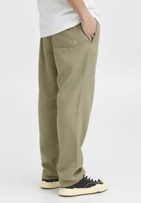 Olivgrüne, strukturierte Hose mit lockerer Passform, seitlichen Taschen und einer Gesäßtasche. Kombiniert mit schwarzen Sneakers mit weißen Schnürsenkeln.