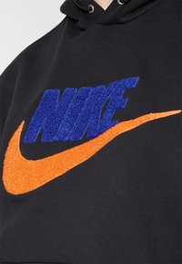 Sudadera negra con letras en relieve en azul "NIKE" y un swoosh naranja. Textura suave con colores contrastantes, enfatizando el diseño de la marca.