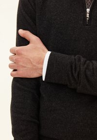 Dunkelgrauer Reißverschluss-Pullover mit gerippten Bündchen und einem kontrastierenden weißen Innencollier. Bemerkenswerte Texturen umfassen eine glatte Oberfläche und gestrickten Stoff.