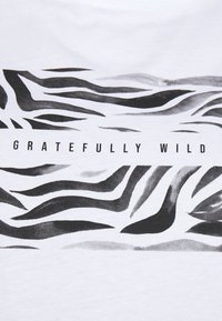 Camiseta blanca con un patrón de rayas de cebra en negro y gris, con el texto "GRATEFULLY WILD" impreso en letras mayúsculas y en negrita.