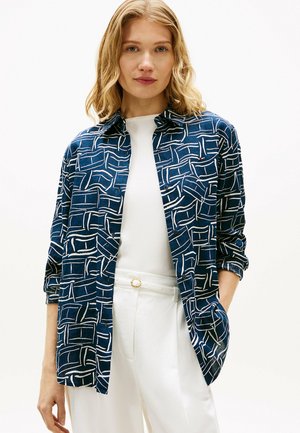 Kvinde iført en blå bluse med lange ærmer og hvide geometriske mønstre over en hvid top samt højtaljede hvide bukser med et bælte med guldlås.