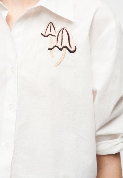 Hvid skjorte med knapper og opkørte manchetter, med broderede pink svampe med sorte omrids på brystområdet.