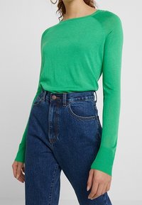 Maglione verde a maniche lunghe con scollo rotondo, dalla texture morbida, abbinato a jeans in denim blu scuro a vita alta con chiusura a bottone.