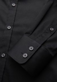 Chemise noire à manches longues avec des boutons argentés sur le poignet et la patte de boutonnage avant, montrant la texture détaillée du tissu et la couture des boutons.