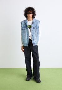 Jeansweste mit zwei Brusttaschen, über einem langärmeligen weißen T-Shirt mit Grafik. Schwarze lässige Hose und schwarze Sportschuhe mit runder Sohle.