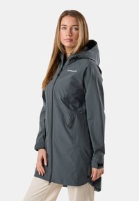 Femme portant une longue veste de pluie à capuche gris foncé avec des poches zippées, debout devant un fond clair uni.