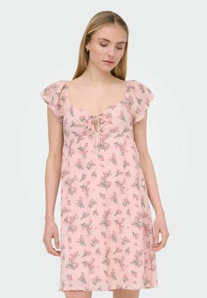 Mladá žena oblečená v ružových kvetovaných šatách s volánovými rukávmi a predným viazaním, pozerá sa dole na jednoduchom svetlom pozadí.