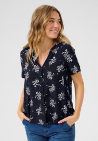 Mörkblå kortärmad blus med V-ringning, prydd med vita blommönster och knappar framtill. Tillverkad av lätt och mjukt material.