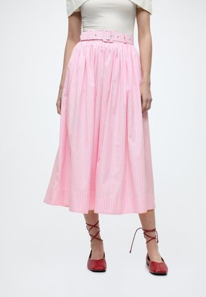 Naine, kes kannab roosat gingham-mustriga midi-seelikut sobiva vööga, valget pluusi ja punaseid pitsilise nöörimisega madalaid kingi, lihtsa taustaga.