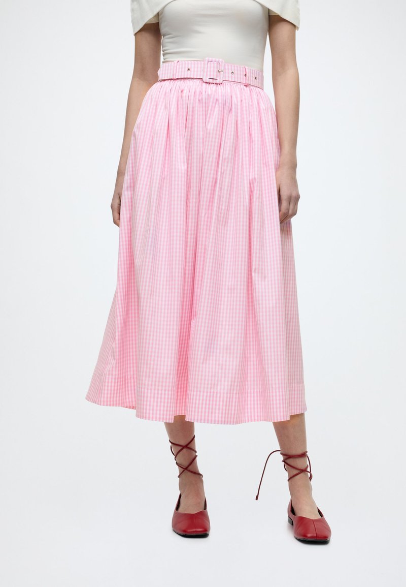 Γυναίκα που φοράει midi φούστα ροζ καρό με ζώνη στο ίδιο ύφασμα, λευκό τοπ και κόκκινα φλατ παπούτσια με κορδόνια, μπροστά σε απλό φόντο.