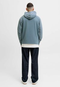 Sweat à capuche bleu clair en tissu texturé, coupe décontractée, superposé sur une chemise blanche, associé à un pantalon en denim foncé et des baskets blanches.