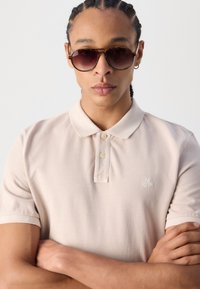 Lys beige polo-skjorte med krage med to knapper, korte ermer, og brodert logo på venstre bryst. Modell med solbriller.
