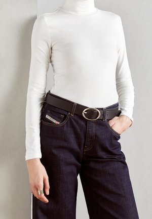 Personne portant un col roulé blanc à manches longues, un jean foncé avec des coutures jaunes, une ceinture noire avec une boucle ovale argentée et une bague argentée à la main gauche.