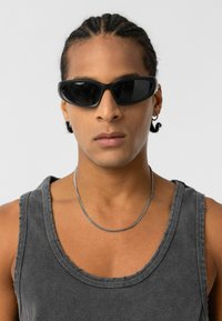 Zwarte zonnebril met een slanke, ovale vorm en een glanzende afwerking. Draagt een grijze tanktop met een ronde halslijn en rafelige randen. Zilveren kettinghalsband.