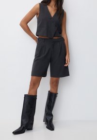 Szary bezrękawnik z guzikami, w połączeniu z szarymi dopasowanymi szortami i wysokimi czarnymi skórzanymi botkami z wydłużonym noskiem i fałdami.