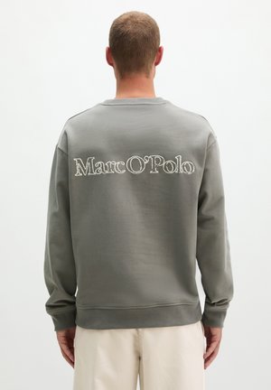 Man draagt een grijze trui met de tekst "Marc O'Polo" op zijn rug, gecombineerd met lichtbeige broek, staande met de rug naar de kijker.