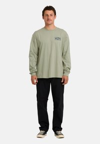 Uomo in piedi che indossa una camicia a maniche lunghe verde salvia con logo sul petto, pantaloni neri e sneakers nere basse con lacci bianchi.