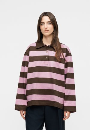 Γυναίκα με μακριά καστανά μαλλιά που φοράει μπλούζα πόλο με μακριά μανίκια, φαρδιές ρίγες σε καφέ και ροζ χρώμα, και σκούρο παντελόνι, στέκεται μπροστά σε ένα φωτεινό φόντο.